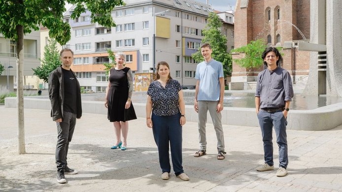 Die Teammitglieder von "ADJUST" stehend im Schatten eines kleinen Baumes vor einem Brunnen
