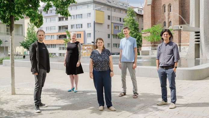 Die Teammitglieder von "ADJUST" stehend im Schatten eines kleinen Baumes vor einem Brunnen
