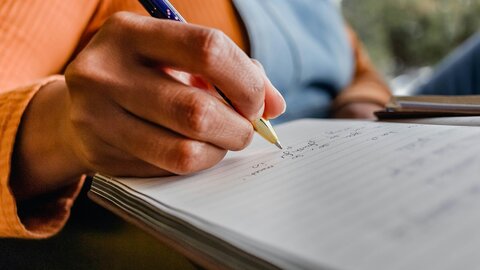 Großaufnahme einer Hand, die mit einem Kugelschreiber Notizen in ein Heft macht