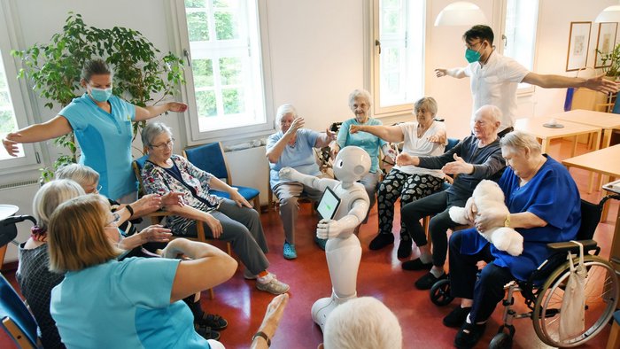 Sitzkreis von &auml;lteren Menschen und Pflegepersonal, in der Mitte ist ein Roboter, der &Uuml;bungen f&uuml;r Beweglichkeit vorzeigt