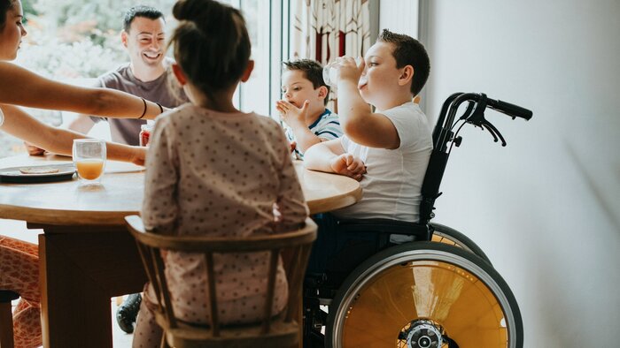 Junge im Rollstuhl sitzt am Esstisch mit seinen Eltern und Geschwistern beim Frühstück