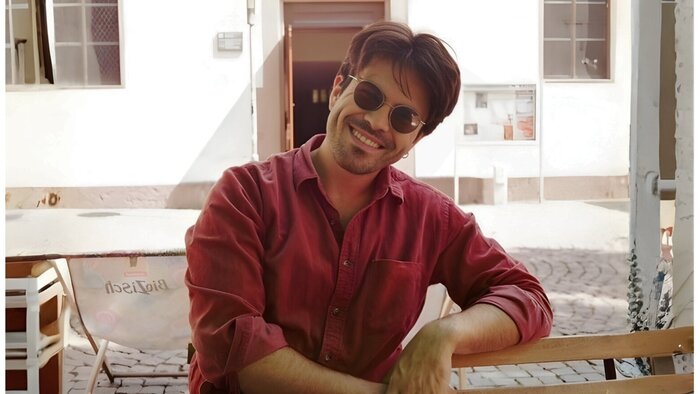 Portr&auml;t eines jungen Forschenden mit Sonnenbrille und rotem Hemd auf einem Platz im Freien