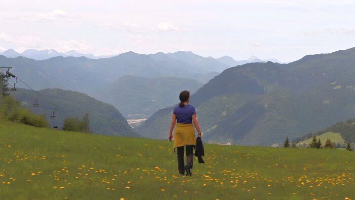 Judith Gl&uuml;ck steht auf einer Blumenwiese in den Bergen und schaut ins Tal und auf das ferne Bergpanorama im Hintergrund