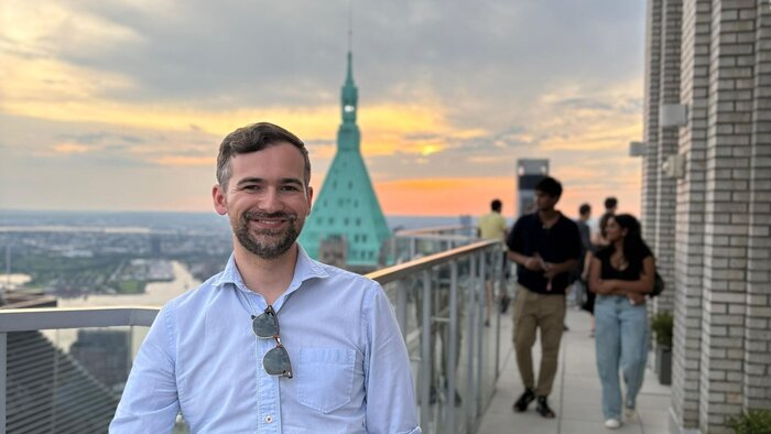 Junger Postdoc in Hemd und Sonnenbrille auf eine Aussichtsplateau, tr&auml;gt Bart und kurze braune Haare, New York im Hintergrund
