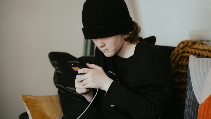 Junge mit schwarzem Kapuzenshirt und schwarzer Mütze sitzt auf dem Sofa und starrt ins Smartphone