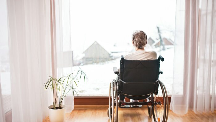 &Auml;ltere Frau sitzt im Rollstuhl vor einem gro&szlig;en Fenster und blickt auf eine verschneite Landschaft