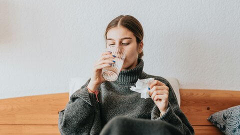 Woman sitting on bed, holding a handkerchief in her left hand and drinking a glass of water
