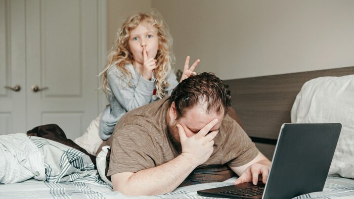 Homeoffice: Vater arbeitet am Laptop im Schlafzimmer mit Tochter auf dem Rücken. Neue Normalität während des Coronalockdowns