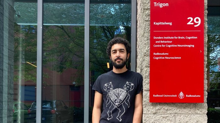 Junger Neurowissenschatler mit dunklen Haaren und Bart, T-Shirt lehnt am Eingang seines Forschungsinstitut neben einer roten Tafel 