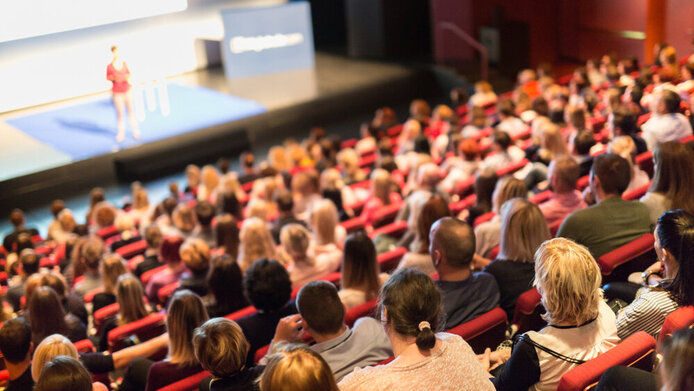 Student:innen in einem großen Hörsaal schauen auf eine Vortragende auf der Bühne
