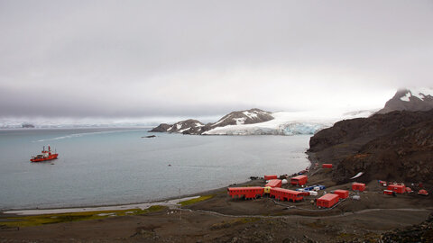 Ein kleines Containerdorf aus roten Hütten direkt an der antarktischen Küste. Im Meer hat ein Polarforschungsschiff angelegt, Schnee und Gletscher im Hintergrund 
