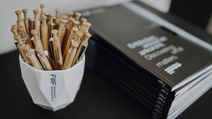 FWF-Kugelschreiber in einer FWF-Tasse neben einem Stapel FWF-Jahresberichten