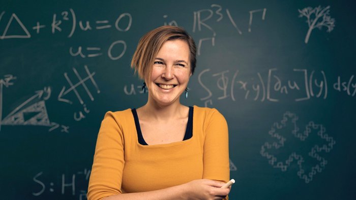 Junge Frau l&auml;chelnd mit orangem T-Shirt und Kreide in der Hand vor einer Tafel mit mathematischen Formeln